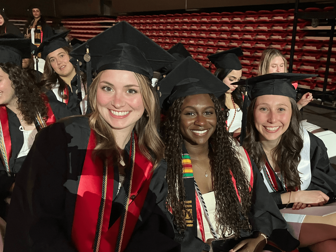three students in graduate cap and gowns smile to the camera