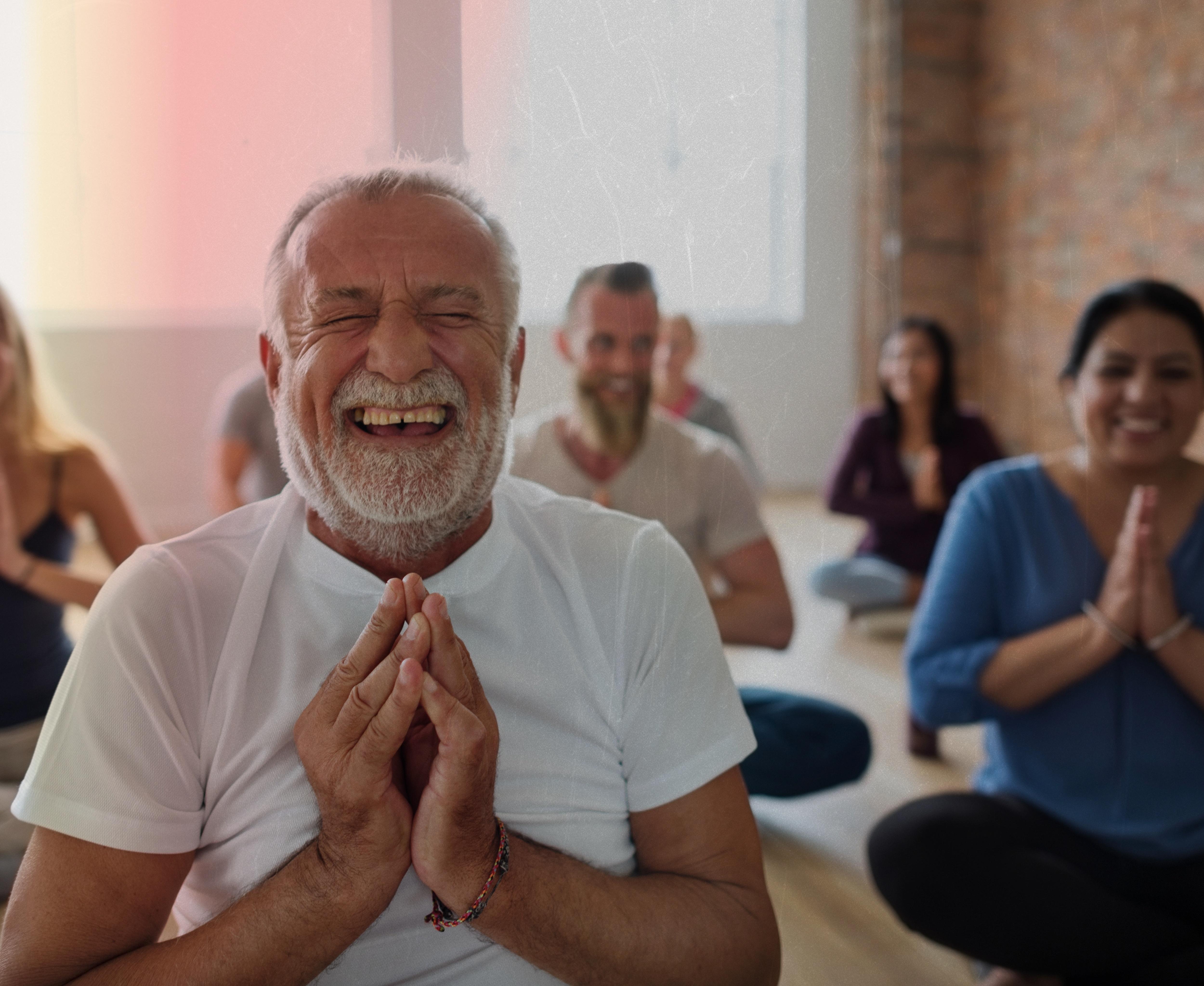 Older man laughs while doing yoga in a room of other people doing yoga