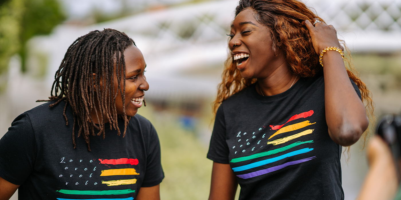 A Black queer couple laughing and looking at each other in t-shirts with rainbow stripes that say Love. 