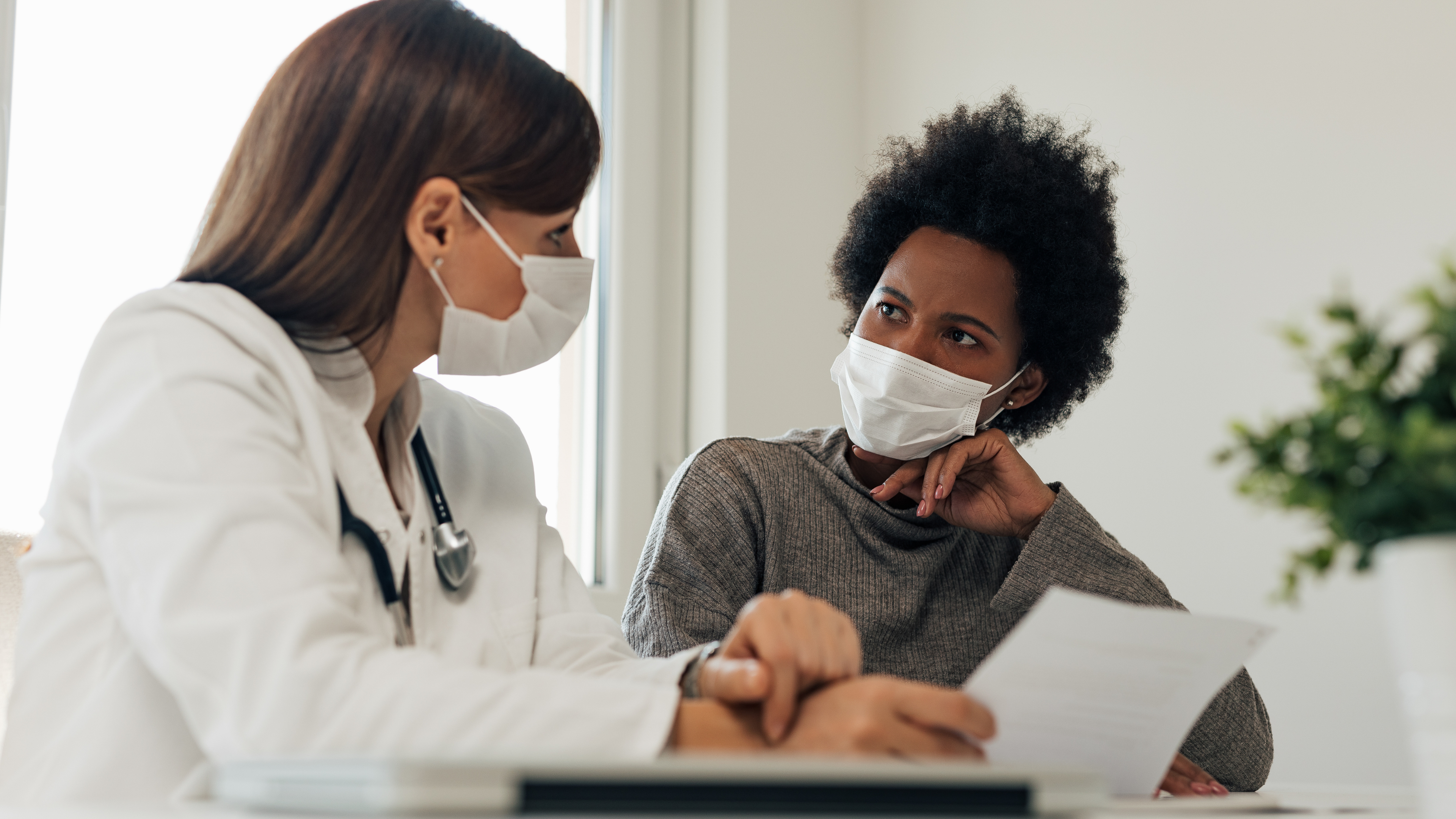 A white female doctor explaining a diagnosis or health information to an African American woman patient, both wearing masks