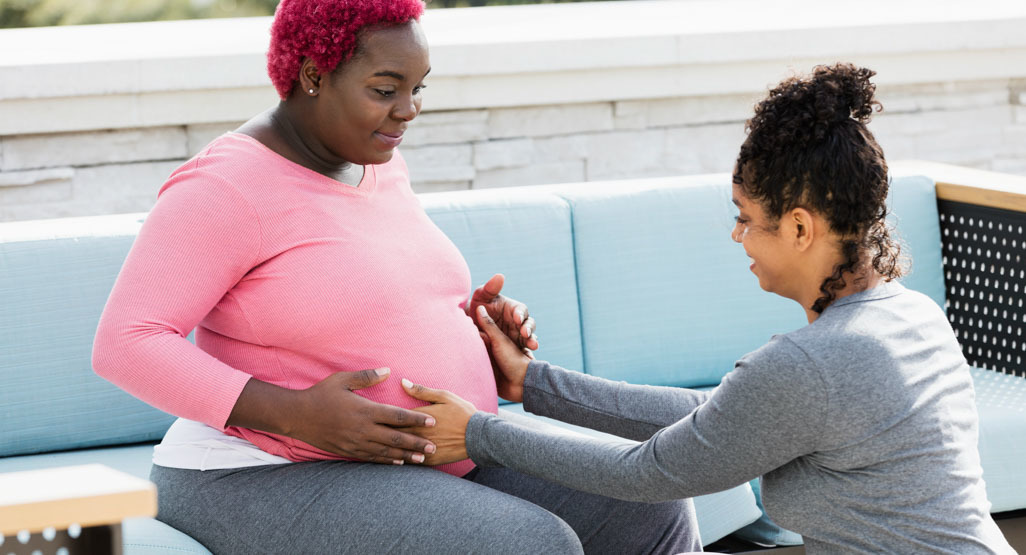 Pregnant Black mom sits on sofa with Black doula putting her hands on the belly