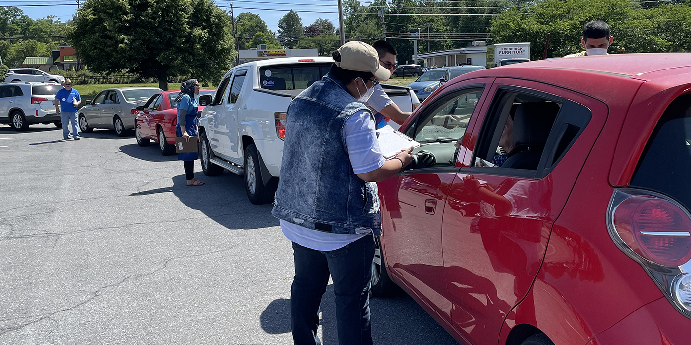 Community health workers from AACF discuss COVID-19 vaccination with residents as they wait at a food distribution event in Frederick in June. A person with a clipboard stands by a red car in a line of cars, talking to the driver.