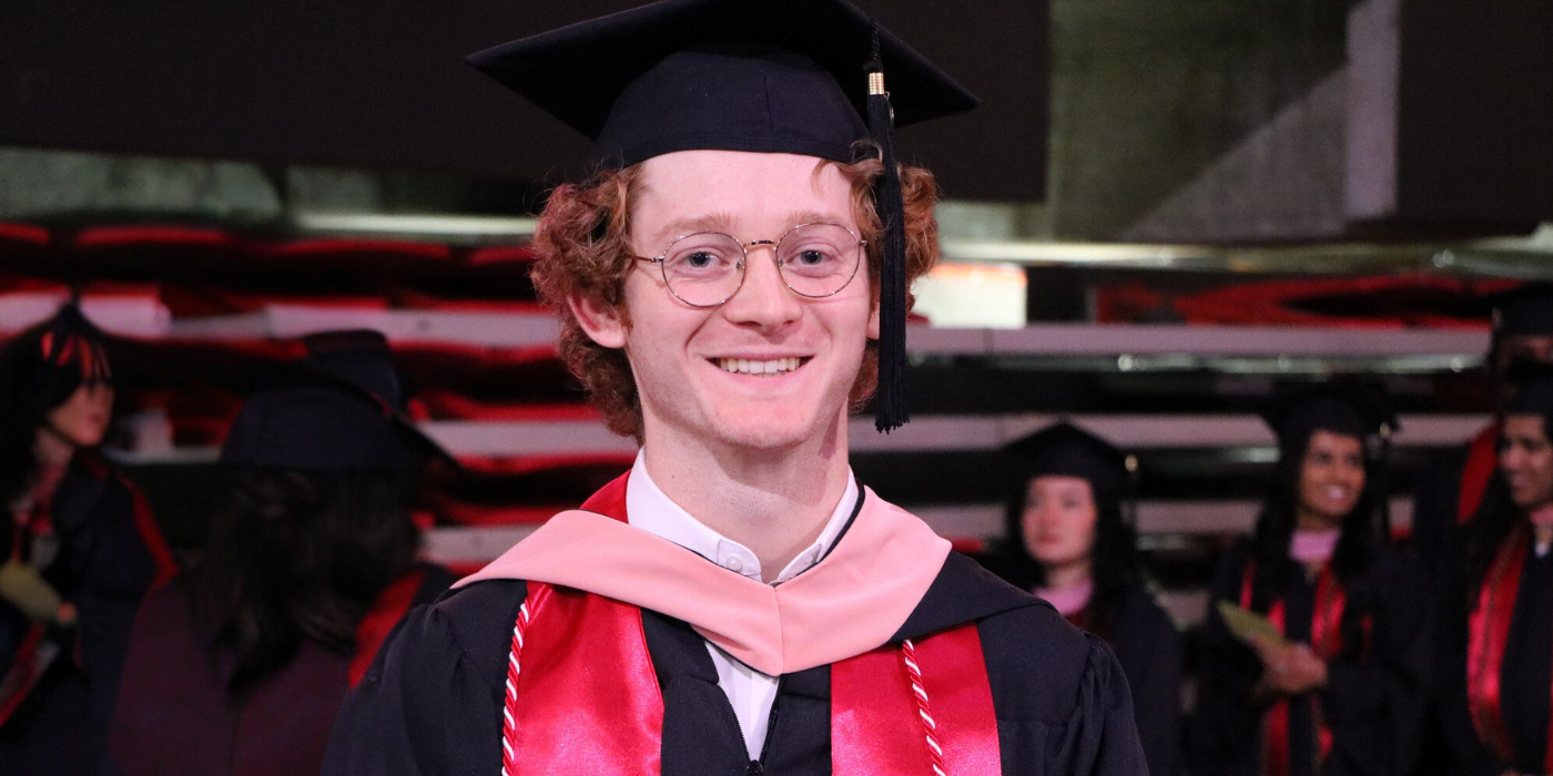 A young white man with red curly hair wearing graduate cap and gown smiles to camera