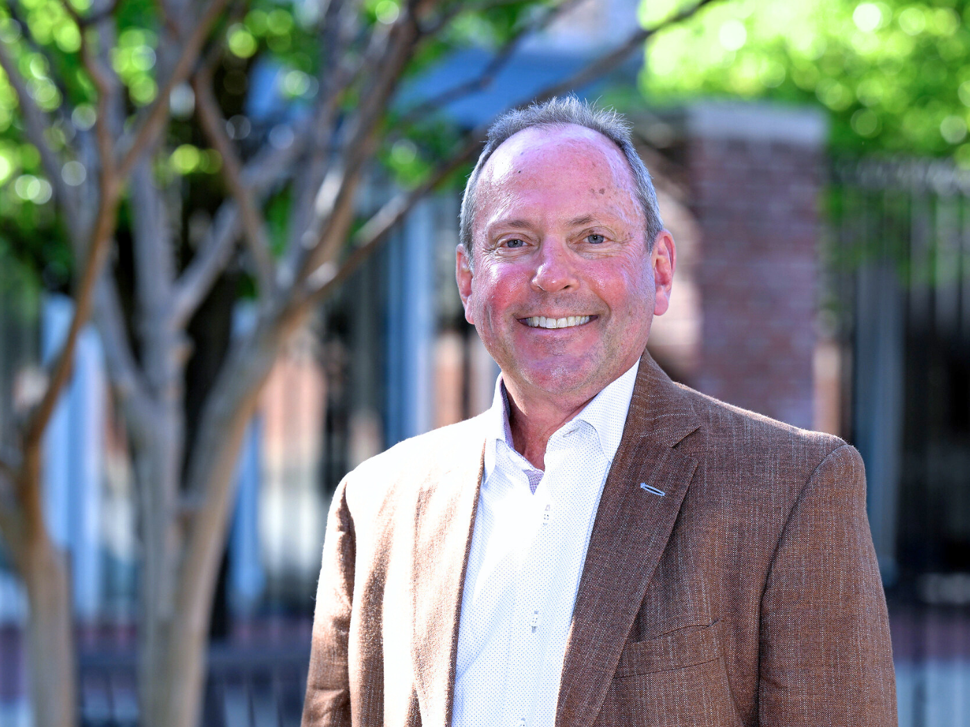 Donor white man in brown suit and white shirt smiles to camera in front of trees