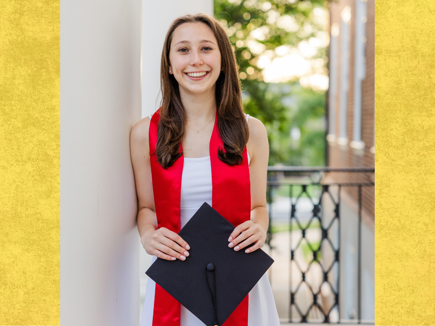 Student in white dress and red graduation scarf, holds black graduation hat, smiles to camera