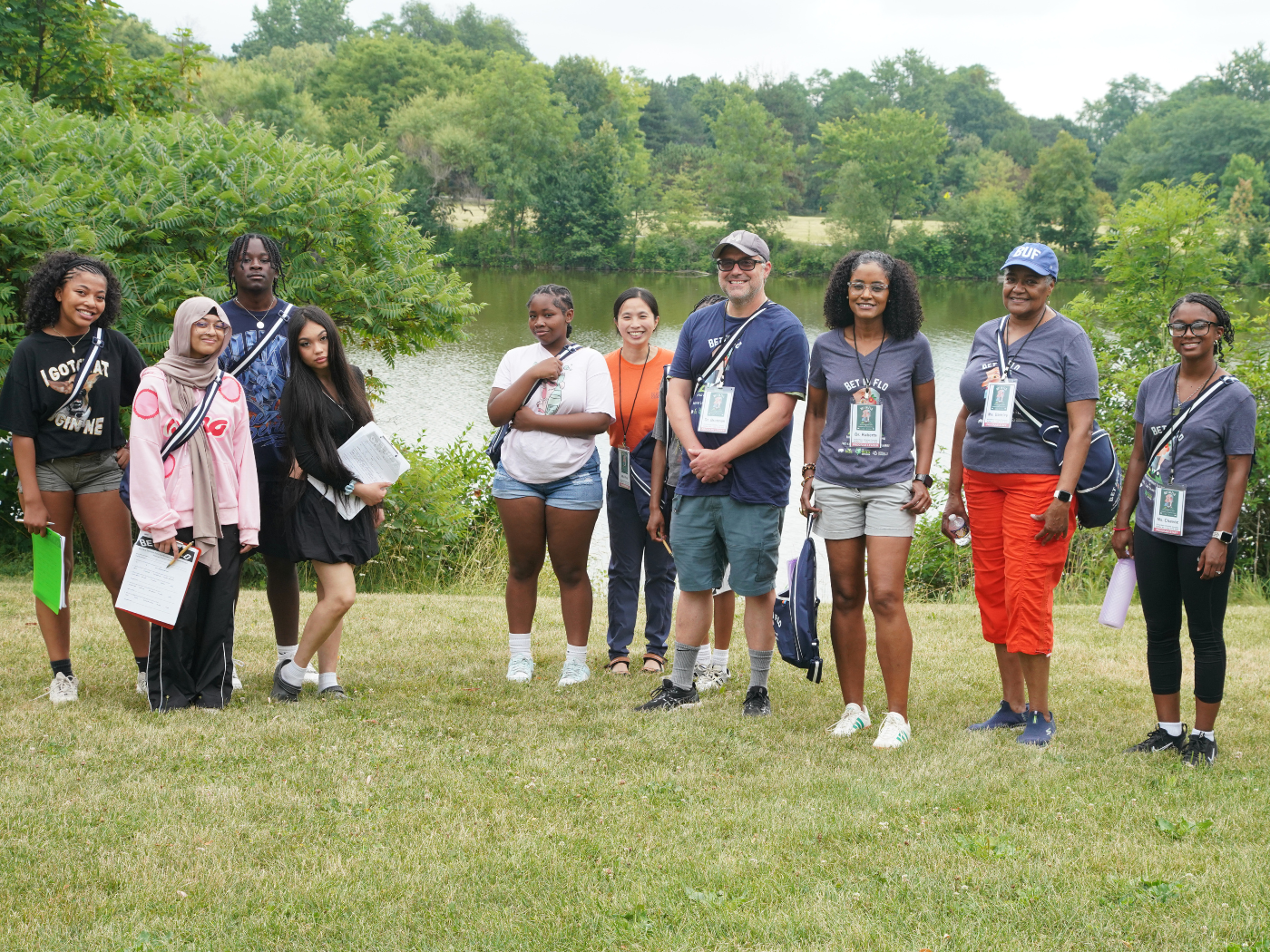 Students and adults stand together smiling to camera in green park area in front of river