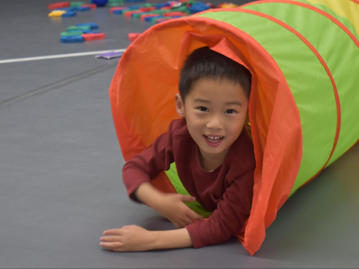 young asian boy in red longsleeve shirt smiles, as he crawls out of a mesh tunnel in the gym