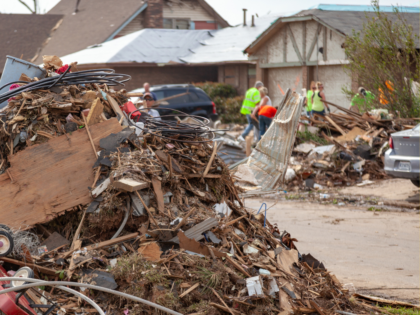 debris of destroyed house is piled up to the left, in background people in neon safety vests clean up