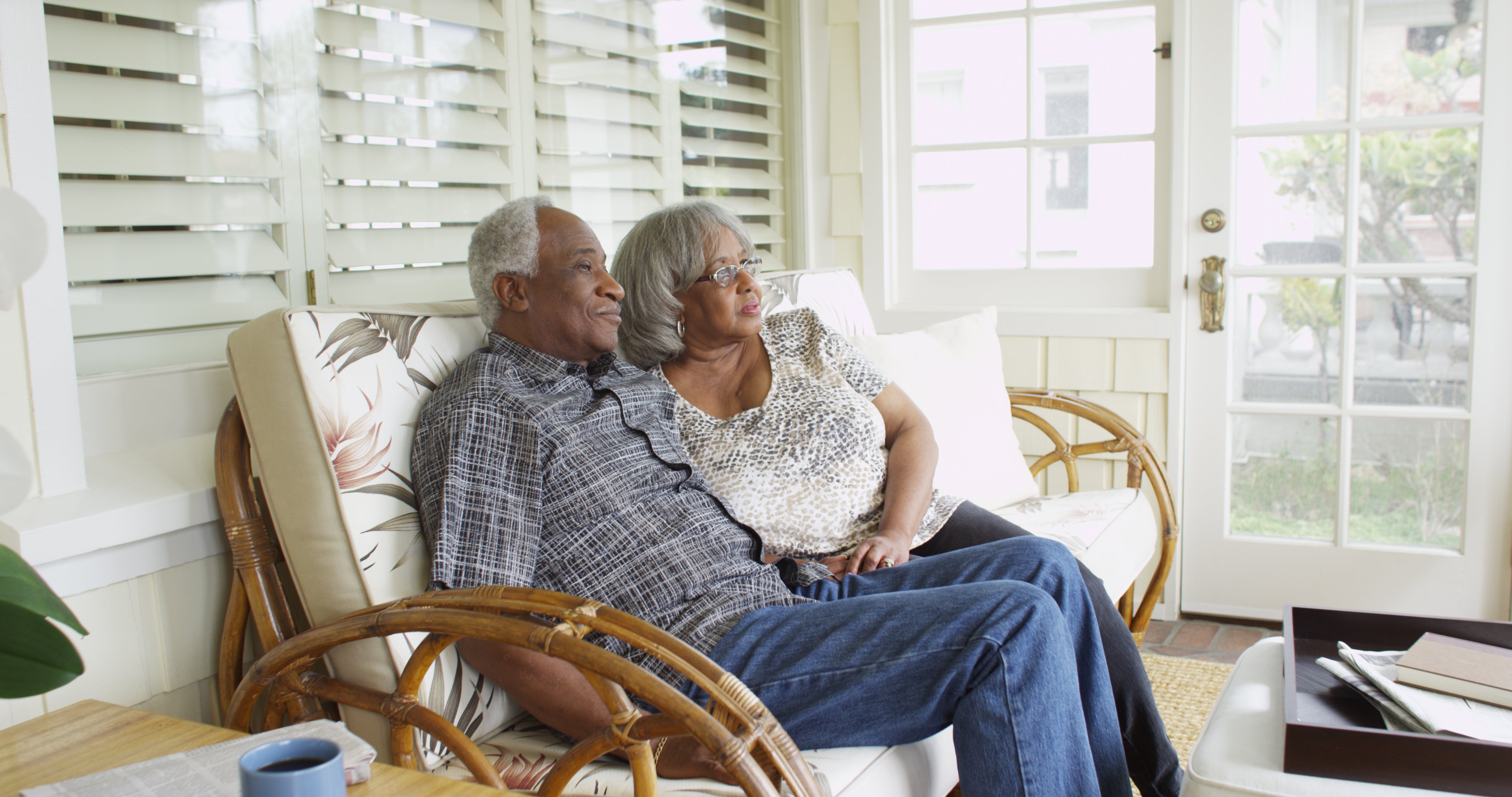 elder black couple man and wife sit on a couch in the sun room of a house, and look out the window