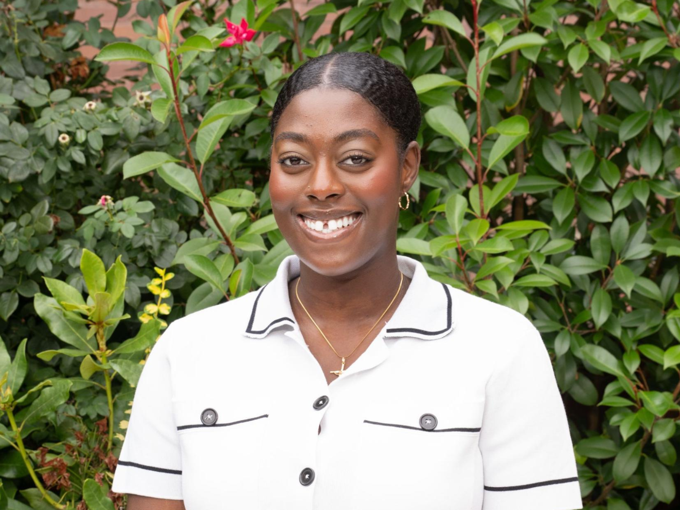 African American student smiles to camera, wearing white shirt in front of green bushes. 