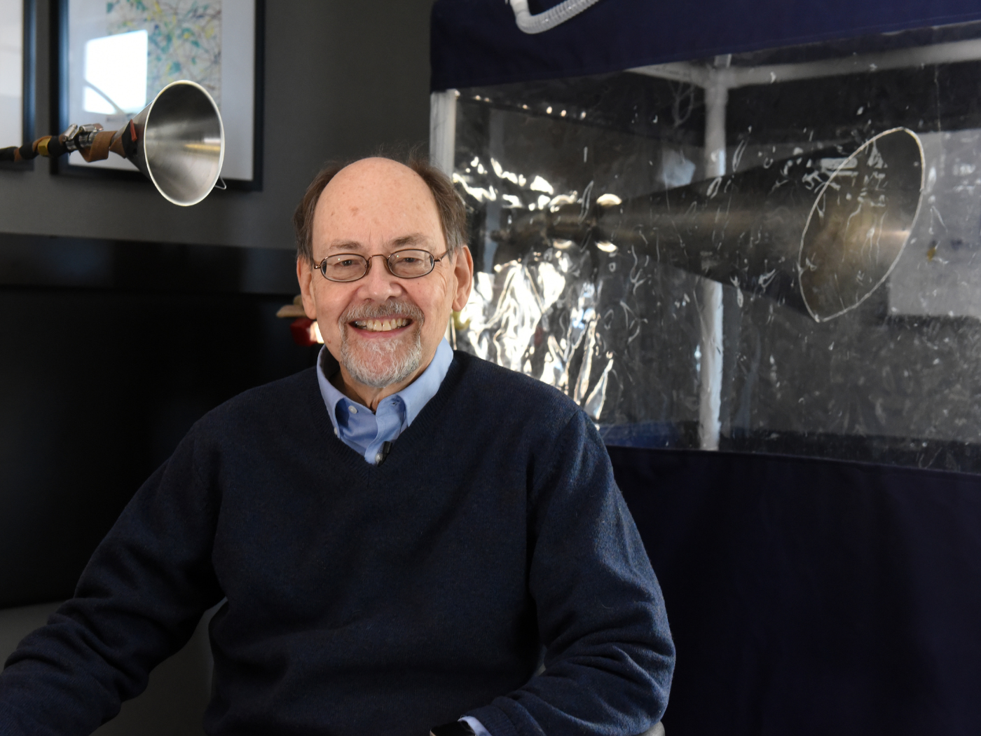 Professor in glasses, wearing blue v-neck and light blue shirt, sits in front of the gesundheit machine, which he invented