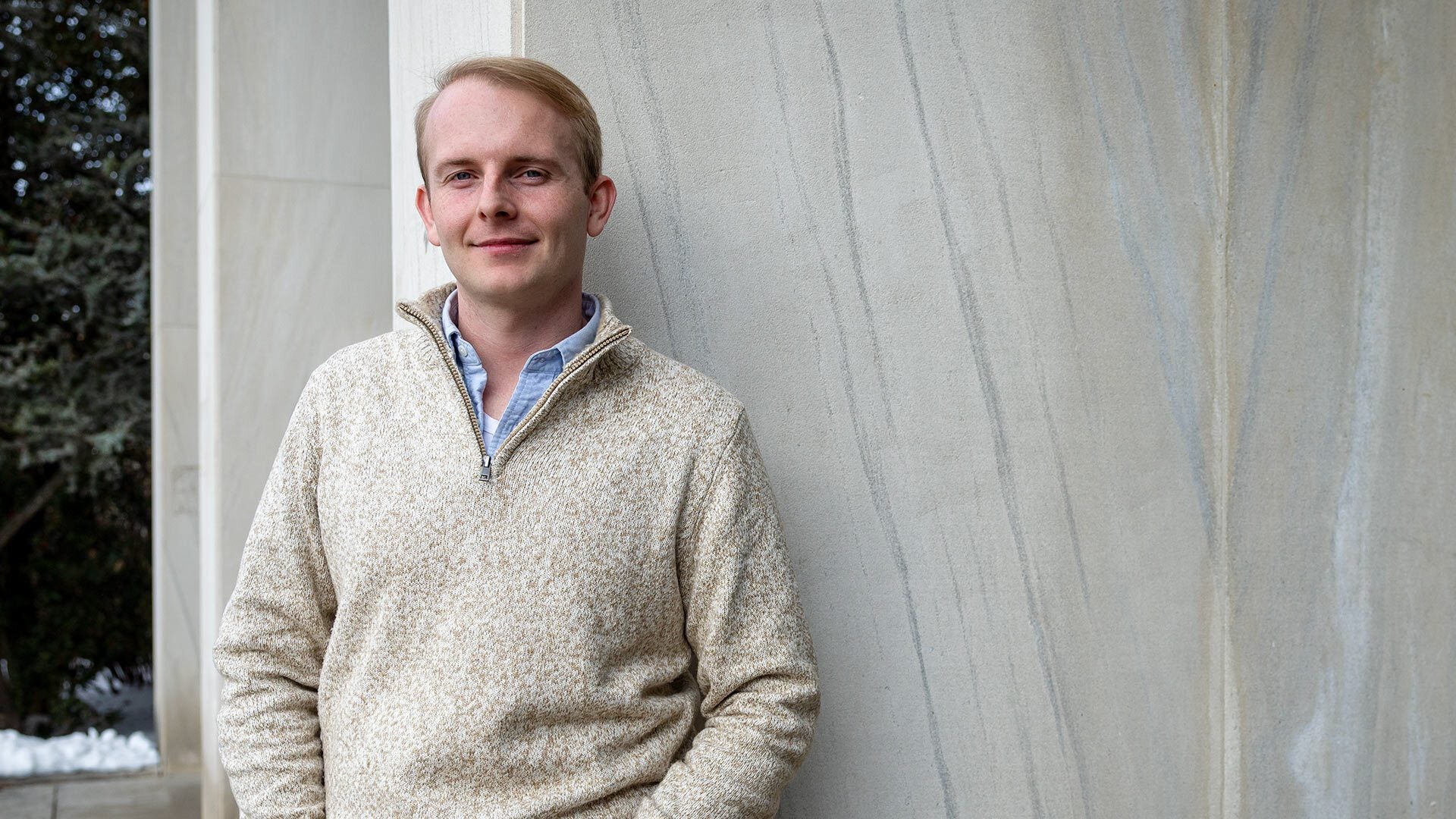 White man with blond hair in beige top standing in front of marble wall looks to camera with a small smile