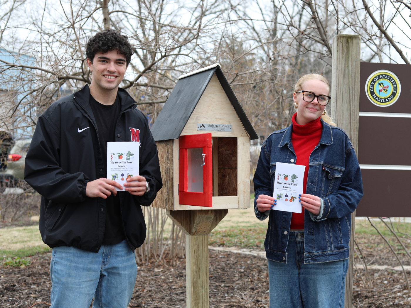 Two students hold recipe book they created, standing in front of a wooden library at Hyattsville Food Forest