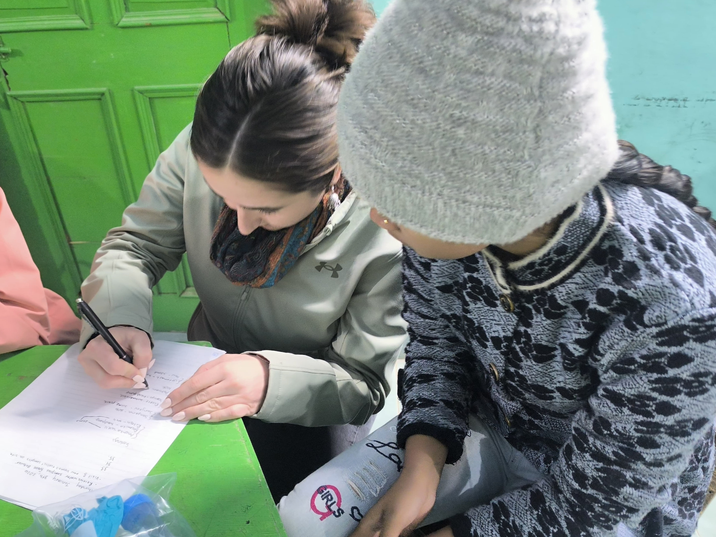 young indian pupil watches as UMD student writes on her paper in a classroom in Varanasi, the table is painted bright green and the walls are light blue