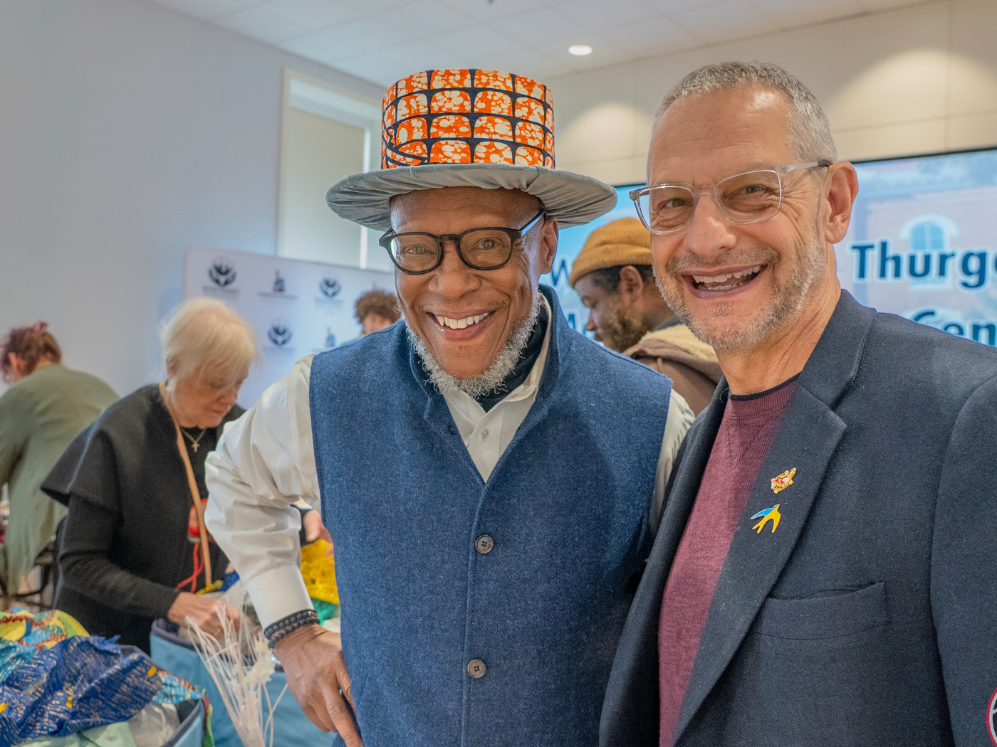 Black male professor wearing top hat made from African fabric poses with white male Dean in a blue sports jacket also smiling