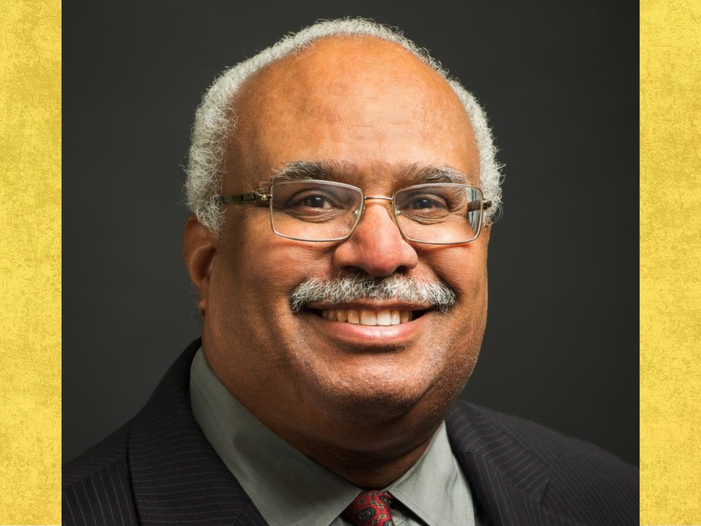 Black older man with greying moustache and rimless spectacle smiles to camera, wearing a dark suit jacket and reddish tie
