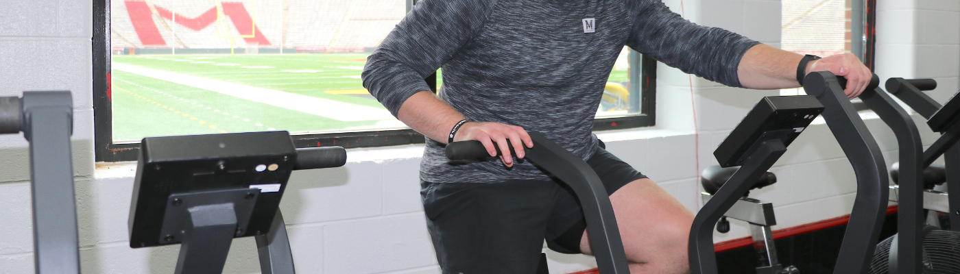 Student on workout equipment in the gym with a view of the football stadium outside the window 