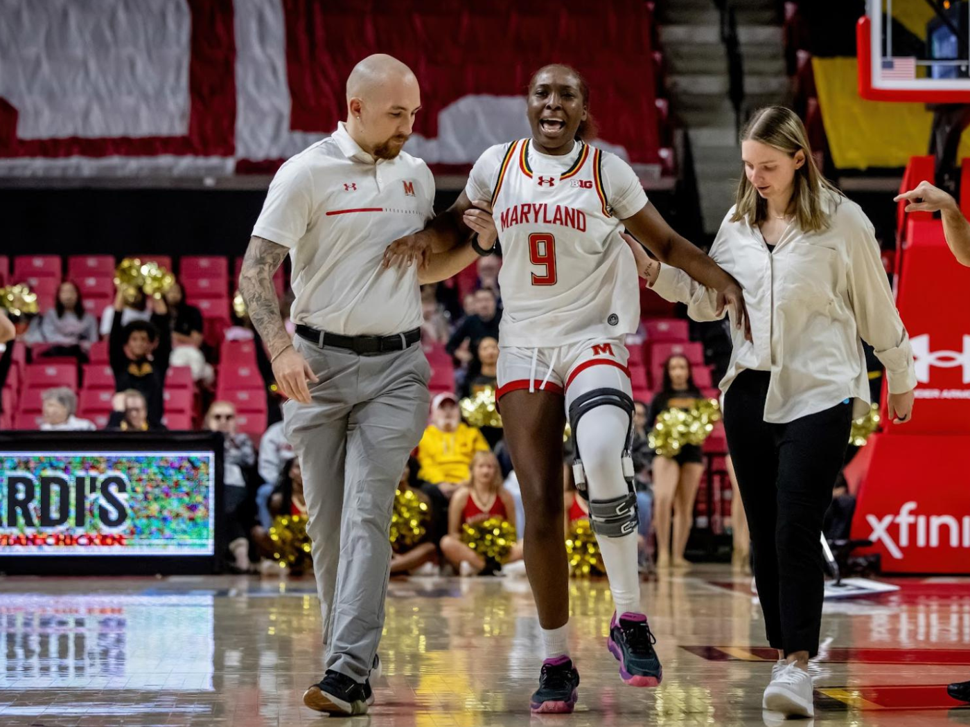 Man in grey trousers and white shirt helps grimacing female basketball player off the court, helped by a woman in black trousers and white shirt, long brown hair, in background seats