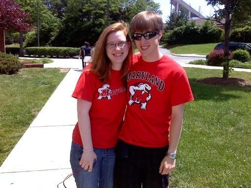 Young white woman and man both wearing red Maryland tshirts pose outside with trees and pathway in background
