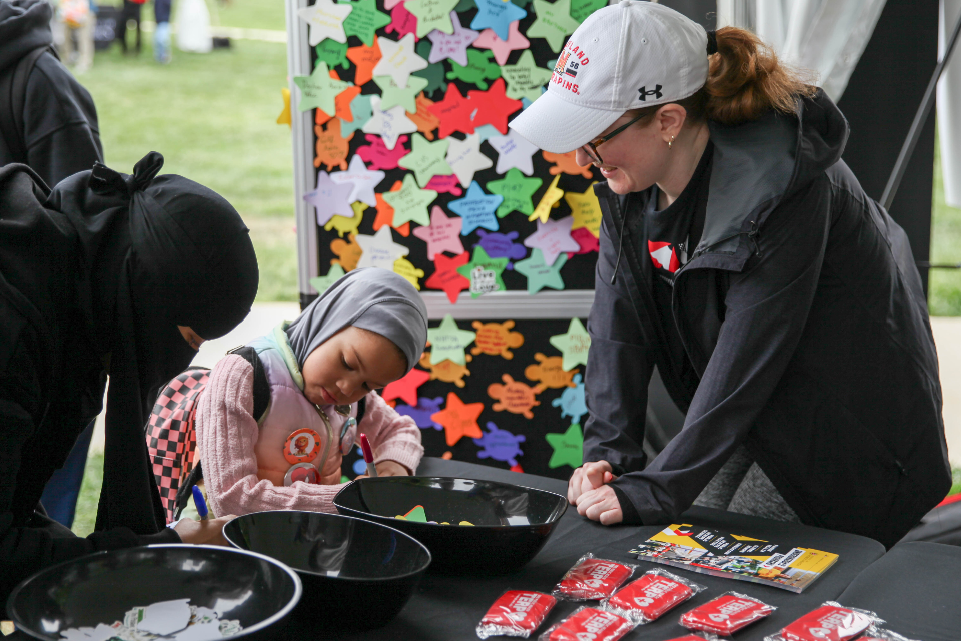 White woman stands behind table on grassy mall smiling, while a woman in black hejabu and young girl with backpack and headdress on draw on paper
