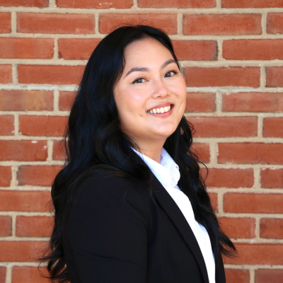 Woman with long black hair in a black blazer and white shirt smiles while standing in front of a brick wall.