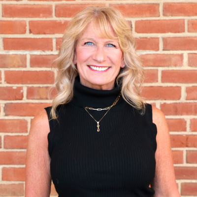 Smiling woman with blonde wavy hair wearing a black sleeveless turtleneck and gold necklace, standing in front of a red brick wall.