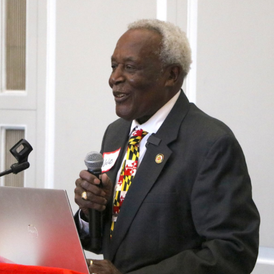 Wesley Queen in a suit speaks into a handheld microphone at a podium with a laptop.