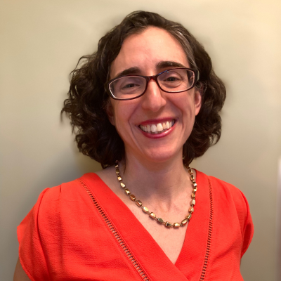 Headshot of a smiling woman with short curly dark hair and glasses, wearing a coral blouse, against a plain light background.