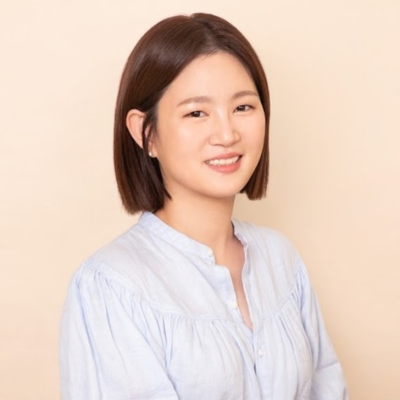 A young woman with straight brown hair in a bob haircut smiling slightly while wearing a light blue blouse against a beige background.
