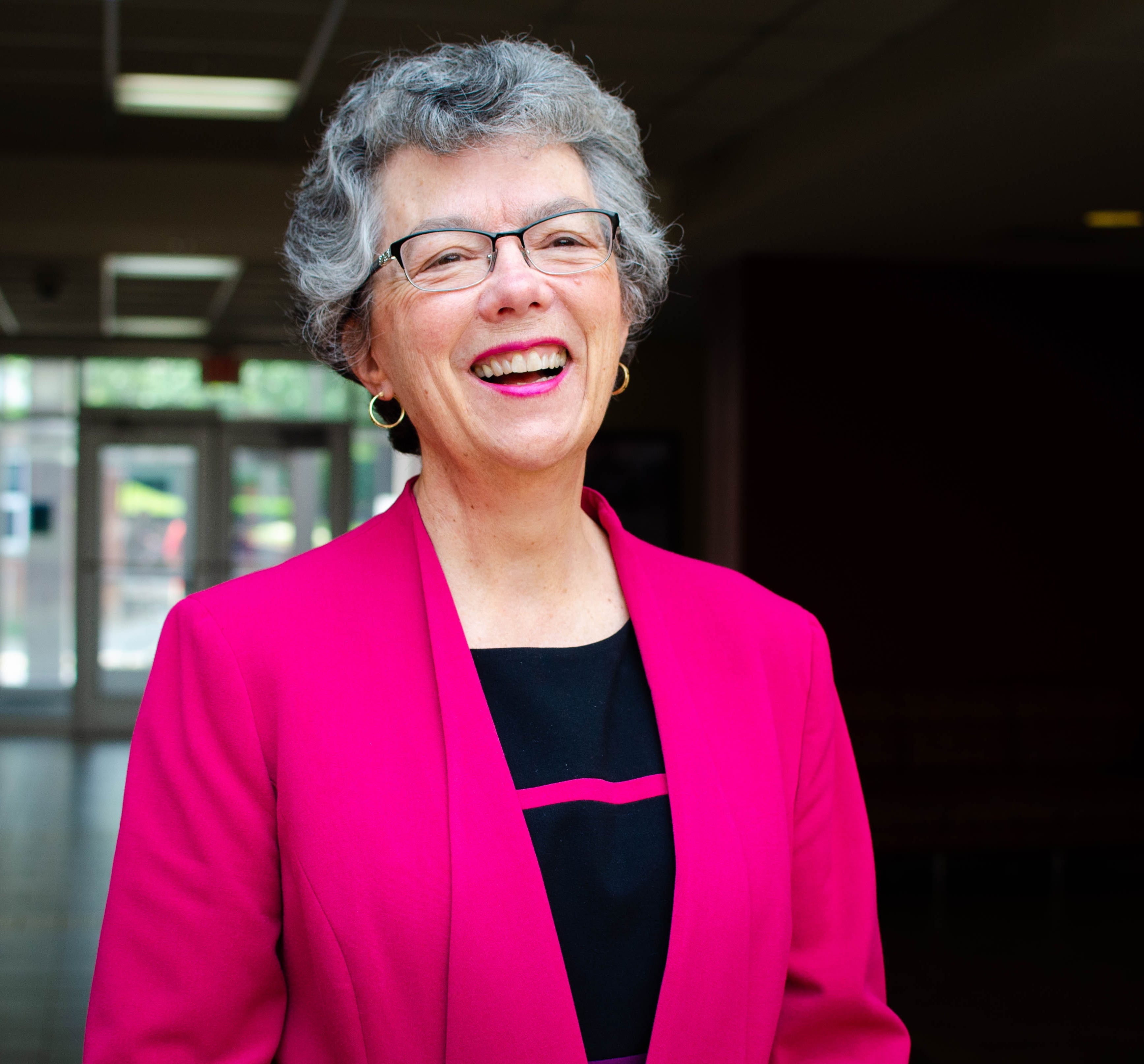 A smiling older woman with short gray curly hair and glasses stands indoors. She is wearing a bright pink blazer over a black top with a pink stripe. The background is softly blurred, showing a hallway with large windows and natural light behind her.