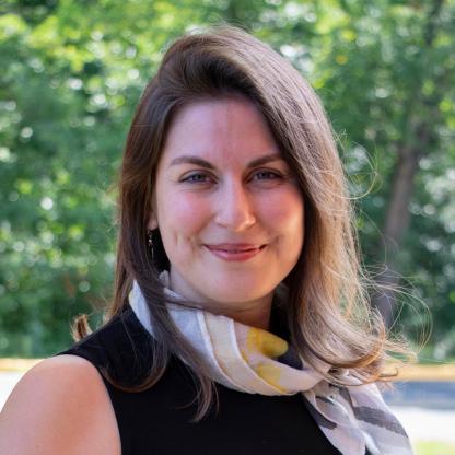 Headshot of a smiling person with long hair wearing a scarf, outdoors with greenery in the background.