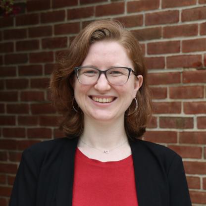 Smiling person wearing glasses stands in front of a brick wall.