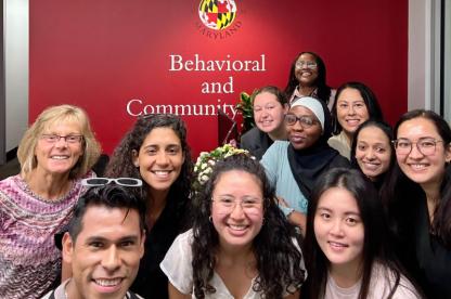 Group of eleven smiling people in front of a red University of Maryland wall that reads “Behavioral and Community Health.