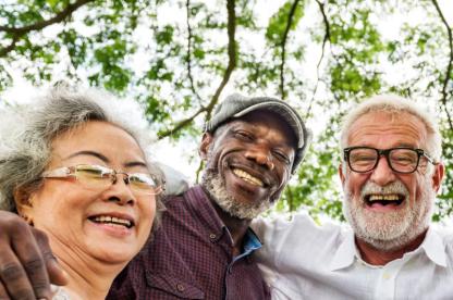 Elders - an asian woman with glasses, a Black man wearing a flatcap and a white-presenting man with glasses — all laughing with arms around each other under tree canopy
