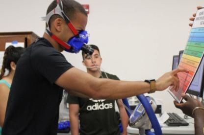A person with respirator on is bicycling and pointing to a chart as another person looks on. 