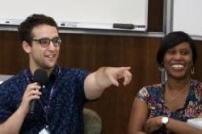 Man holding a microphone points toward the audience while a woman seated beside him smiles.