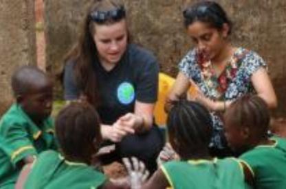 Two women sit and interact with a group of children wearing green uniforms outdoors.