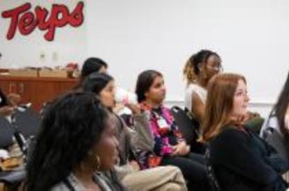 Students sit in an audience during a University of Maryland event, with a Terps logo visible on the wall.