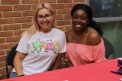 Two women smile while seated together at a table in front of a brick wall.