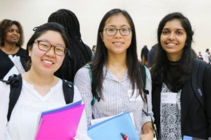 Three students smile at the camera while holding folders and wearing backpacks at a campus event.