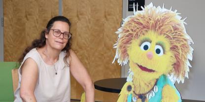 woman with long brown hair and glasses posing next to a cutout of a Muppet that she helped design. Important text includes event title, Kami's Impact: When Sesame Street Took on HIV/AIDS, along with event date of September 23, 2006, the location of Hornbake and time of 3:30 PM