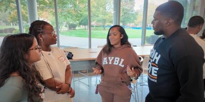 Four students of colour stand together, three listen to one student speaking, who wears beige sweatshirt and pants, in a room with floor to ceiling windows, trees, grass and pathways are seen in background