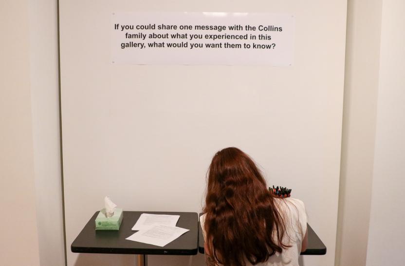 A while wall with a question written on it, beneath the words is a table with paper and pencils, and a woman with long brown hair is seen from behind sat at the table. 
