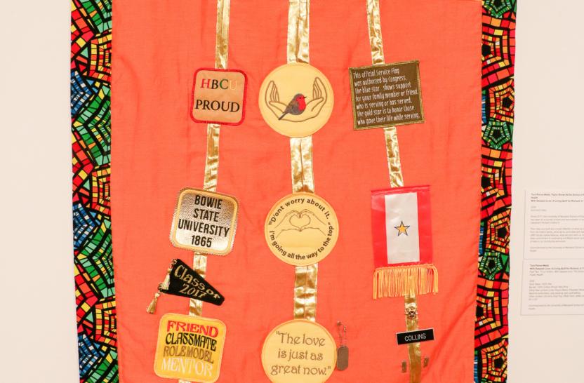 Part of a quilt with lining of African fabric, orange fabric background with symbols representing the life of Lt. Collins, hanging on white wall of gallery.