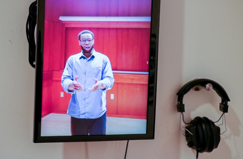 On a screen a Black man sings with arms outstretched; next to screen a pair of headphones hangs on wall. 