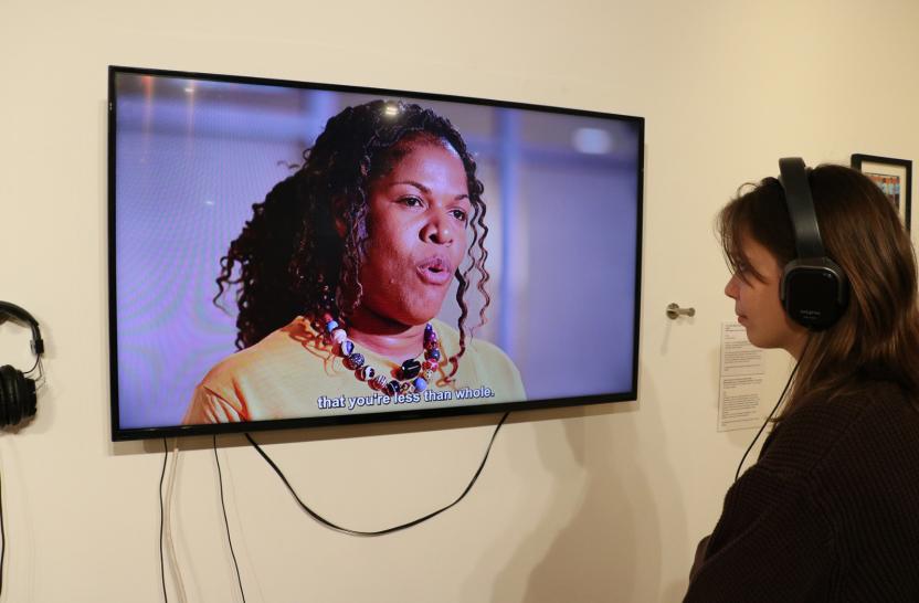 Video on wall shows Black woman talking, with young white woman wearing headphones, listening. 