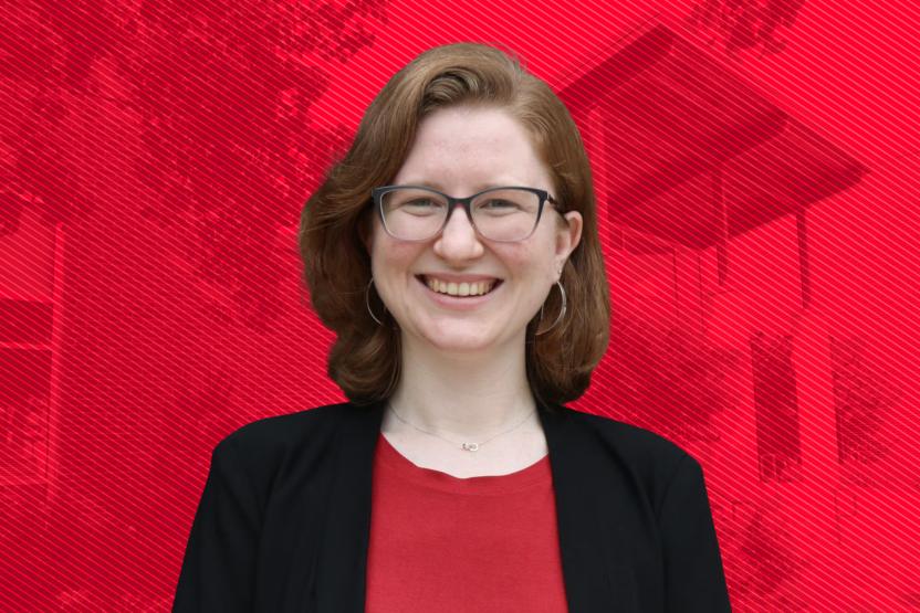 A smiling woman with glasses and shoulder-length brown hair, wearing a black blazer and red top, posed against a bright red geometric background.