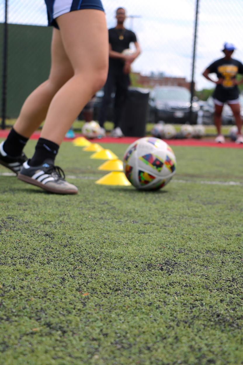 Soccer ball in front of a soccer player's legs with cones in the background.