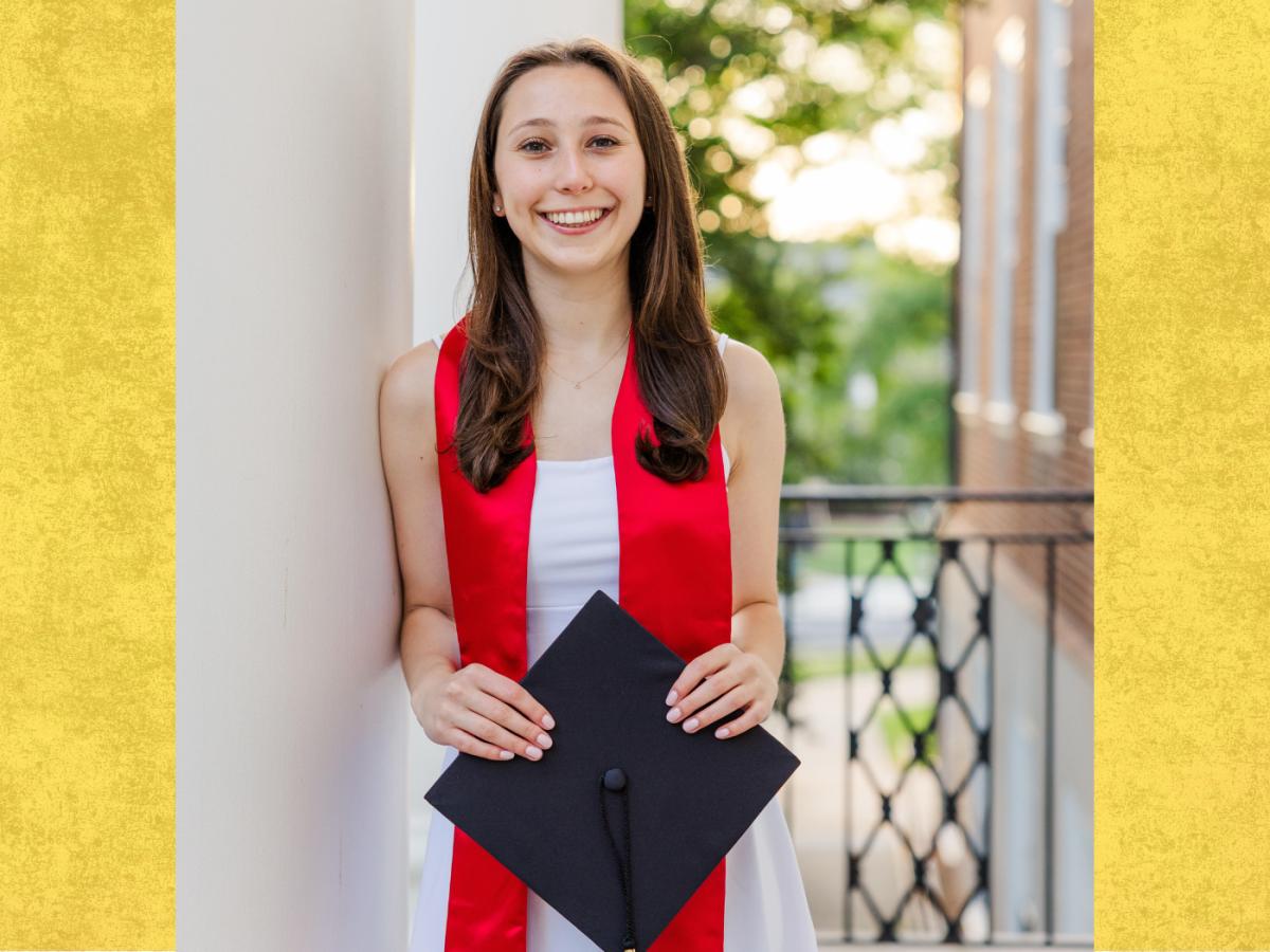 Student in white dress and red graduation scarf, holds black graduation hat, smiles to camera