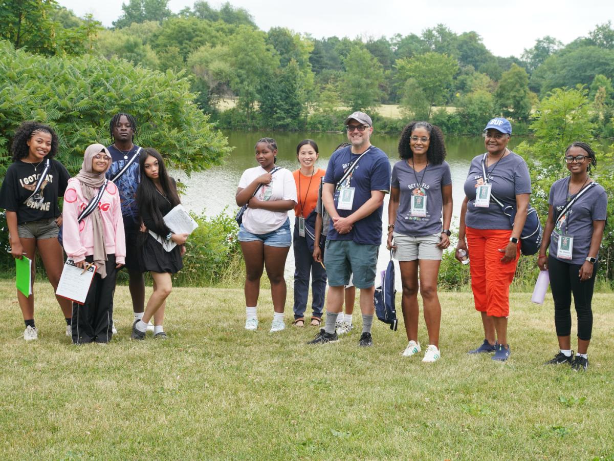Students and adults stand together smiling to camera in green park area in front of river